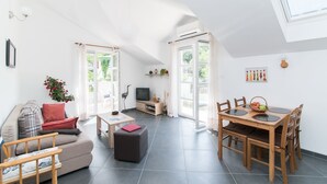 Un vaste espace de vie doté d’un haut plafond, une table à manger avec des chaises, un canapé avec des coussins, une petite table avec un coussin rouge et un téléviseur.