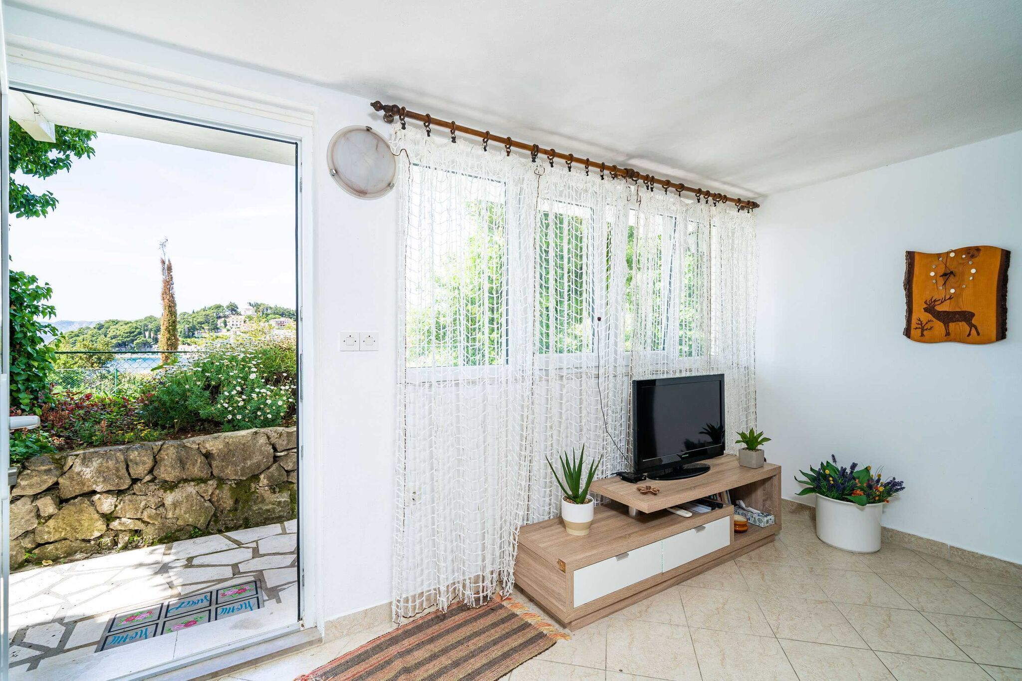 Een woonkamer met een televisie op een houten dressoir, een kamerplant en uitzicht op groen.