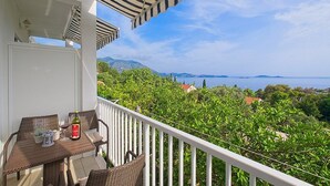 A balcony with wicker furniture, a table with a bottle and glasses, and a view of the sea and greenery.