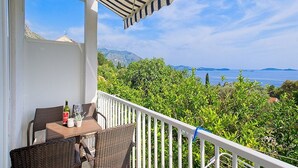 A balcony with wicker furniture, a bottle, and a glass on a table, overlooking a scenic view of a body of water and greenery. - Apartments Grljević - Studio Apartment with Sea View and Balcony (A2) (Mlini)