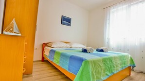Un dormitorio con cama, armario de madera, una estantería pequeña en la pared y ventana con cortinas.