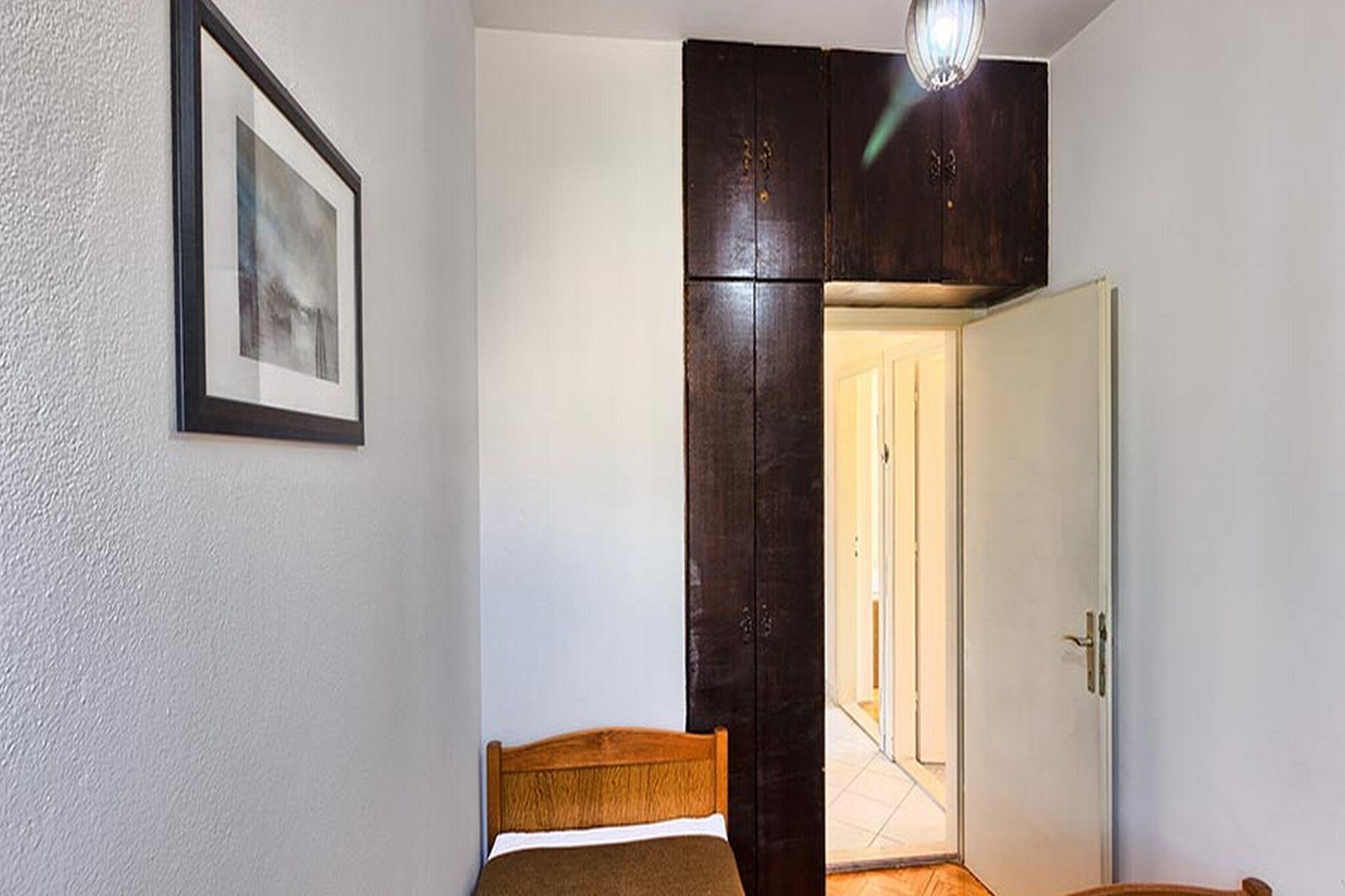 A bedroom with a bed, a framed picture on the wall, a wardrobe, and a door leading to another room.