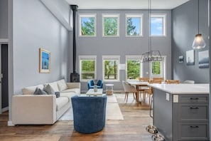 Una sala moderna con sofá, un sillón azul, un comedor con mesa y sillas, y una cocina con armarios y una lámpara colgante.