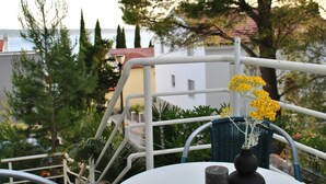 A balcony with a white railing, a table with a vase of yellow flowers, and a view of the sea and buildings in the background. - Apartments Smokvica - One Bedroom Apartment (Starigrad-Paklenica)