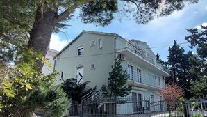 A two-story building with a balcony, surrounded by trees and a metal fence. - Apartments Smokvica - One Bedroom Apartment (Starigrad-Paklenica)