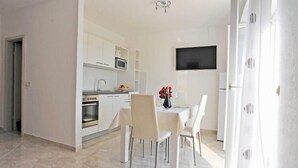 A modern kitchen with white cabinets, a microwave, and an oven. There is a dining table with chairs and a vase of red flowers. A flat-screen TV is mounted on the wall. - Apartments Lenka - One Bedroom Apartment with Balcony and Center View A3 (Sutivan)