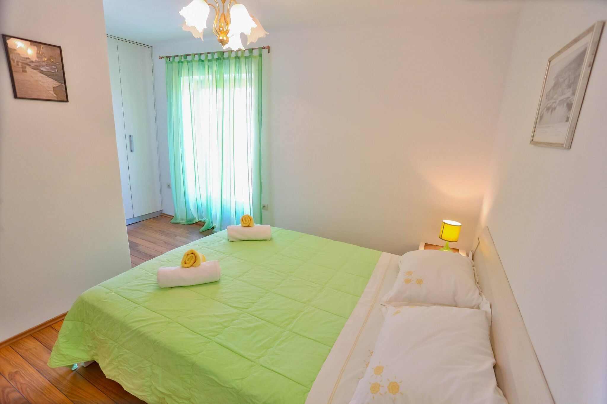 A neatly made bed with green bedding and white pillows, a bedside lamp, and a view of a hallway with a door and curtains.