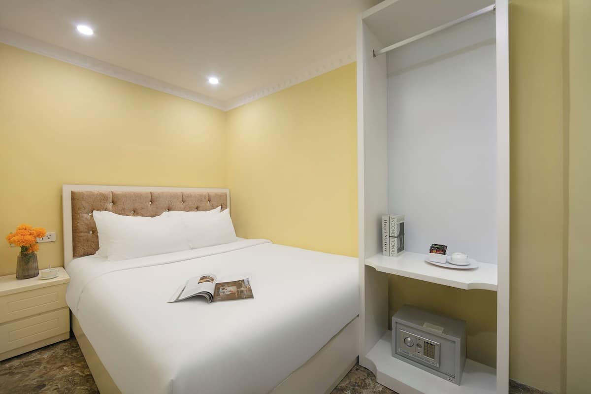 A hotel room with a bed, a nightstand with a vase of flowers, a shelf with a radio, and a white wardrobe.