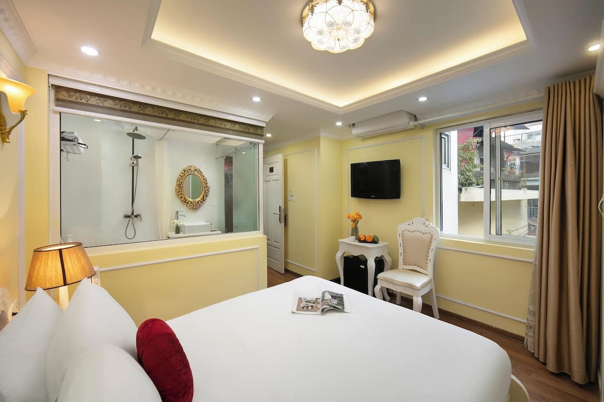 A hotel room with a bed, a bedside table with a magazine, a chair, a TV, and a window with curtains.