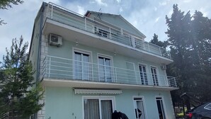 Un bâtiment à deux étages doté d’un balcon, avec une voiture garée devant et une personne qui travaille près de l’entrée.