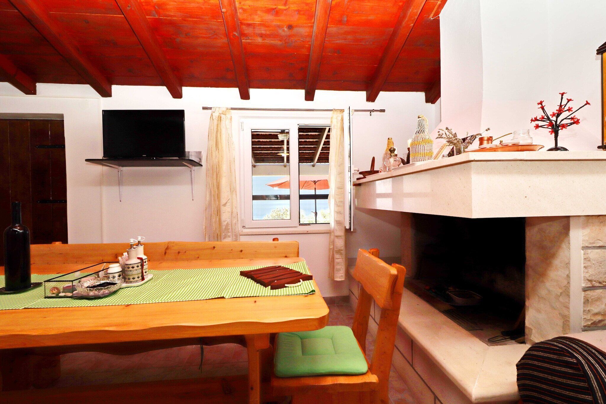 A wooden dining table with a green placemat, a wooden chair with a green cushion, a fireplace, and a television mounted on the wall.