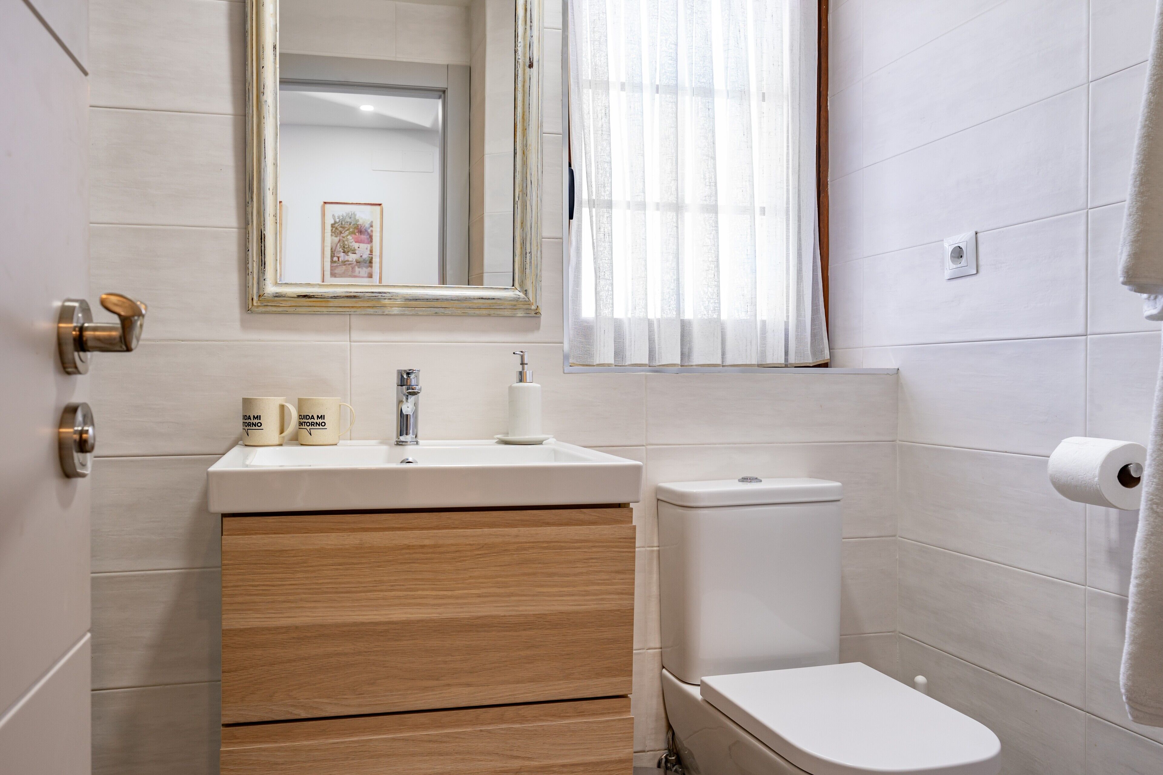 A bathroom with a sink, mirror, toilet, and a window with curtains.