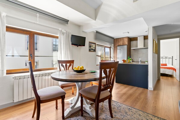Un coin repas comprenant une table ronde, des chaises, un téléviseur fixé au mur, ainsi qu’un espace cuisine avec un réfrigérateur et un évier.
