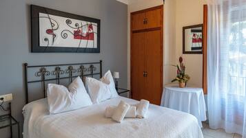 A neatly made bed with white linens and pillows, a wooden door, a small table with a vase of flowers, and a framed picture on the wall.
