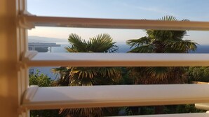 Une vue à travers les stores d’une fenêtre, donnant sur une zone côtière avec des palmiers et un pont au loin.