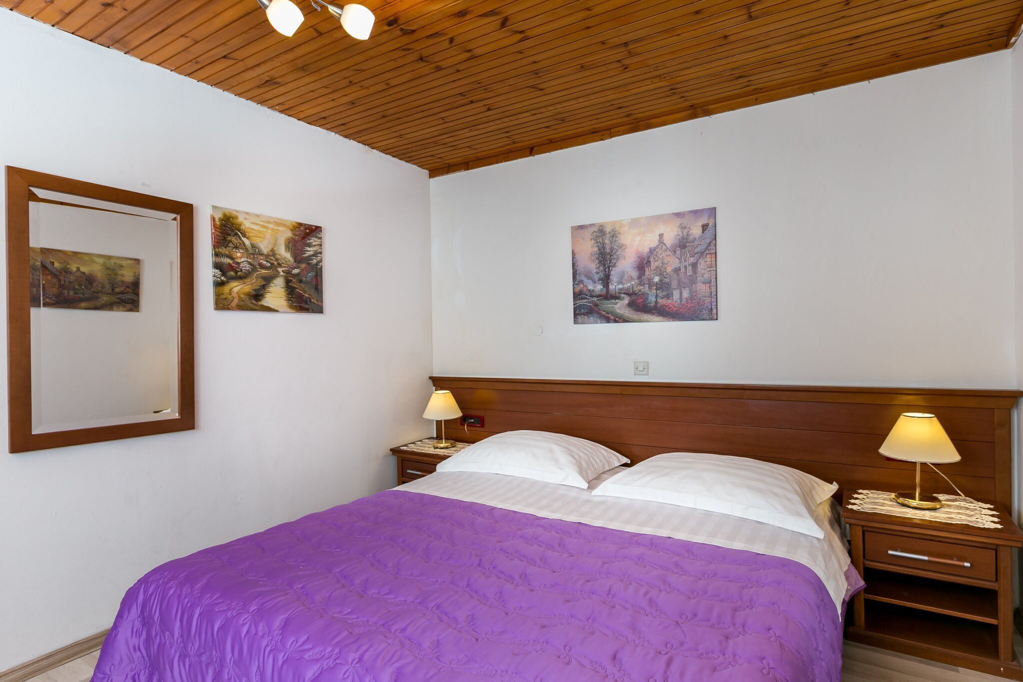 A bedroom with a wooden ceiling, a bed with a purple bedspread, two bedside lamps, and three paintings on the wall.