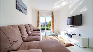 A modern living room with a sofa, a glass coffee table, a flat-screen TV, and a balcony with curtains.