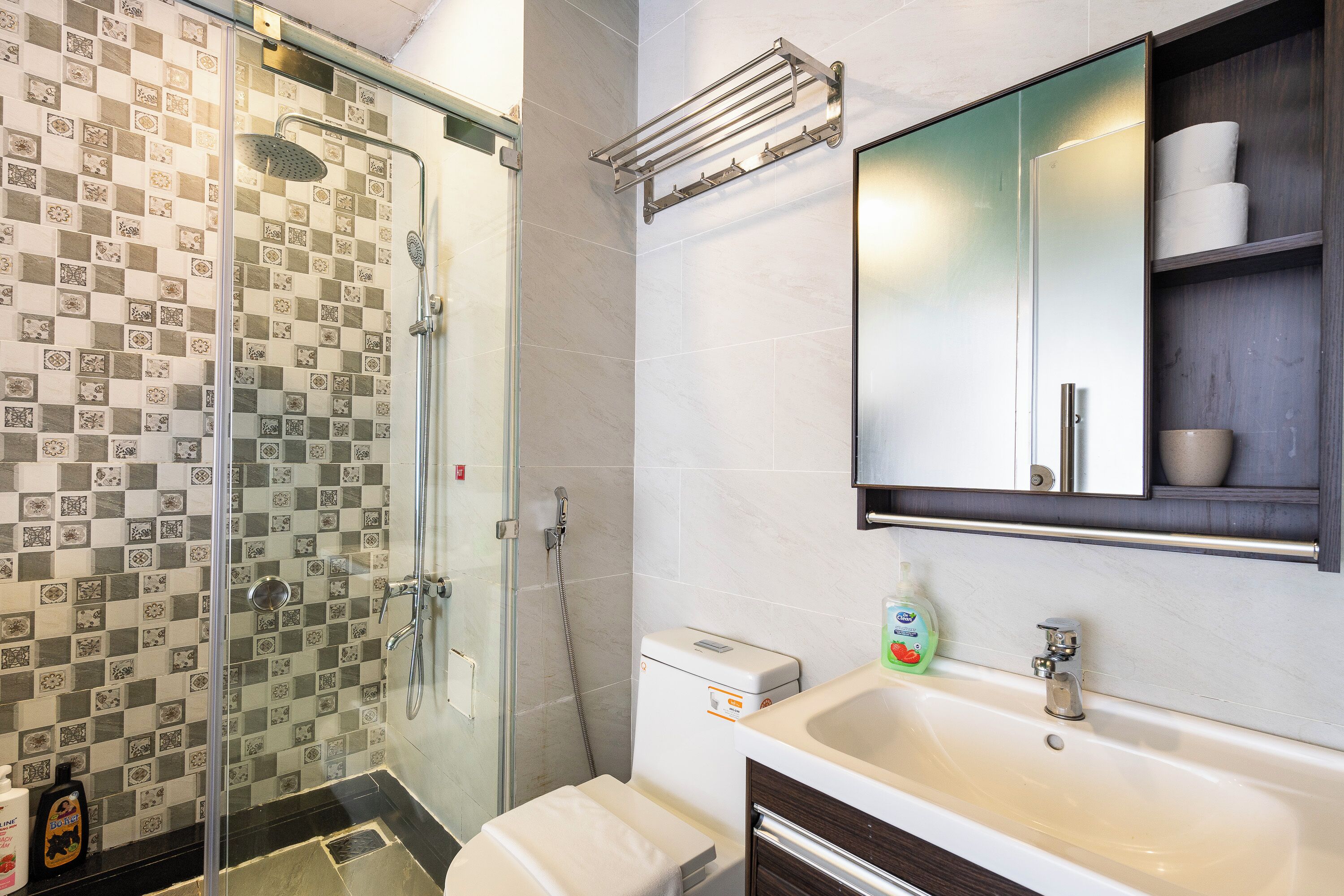 A bathroom with a glass shower enclosure, a toilet, a sink with a mirror above it, and a shelf with toiletries.