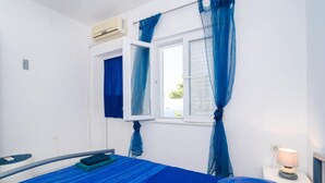 Un dormitorio con cama, una ventana con cortinas azules, una lámpara de noche y un equipo de aire acondicionado.