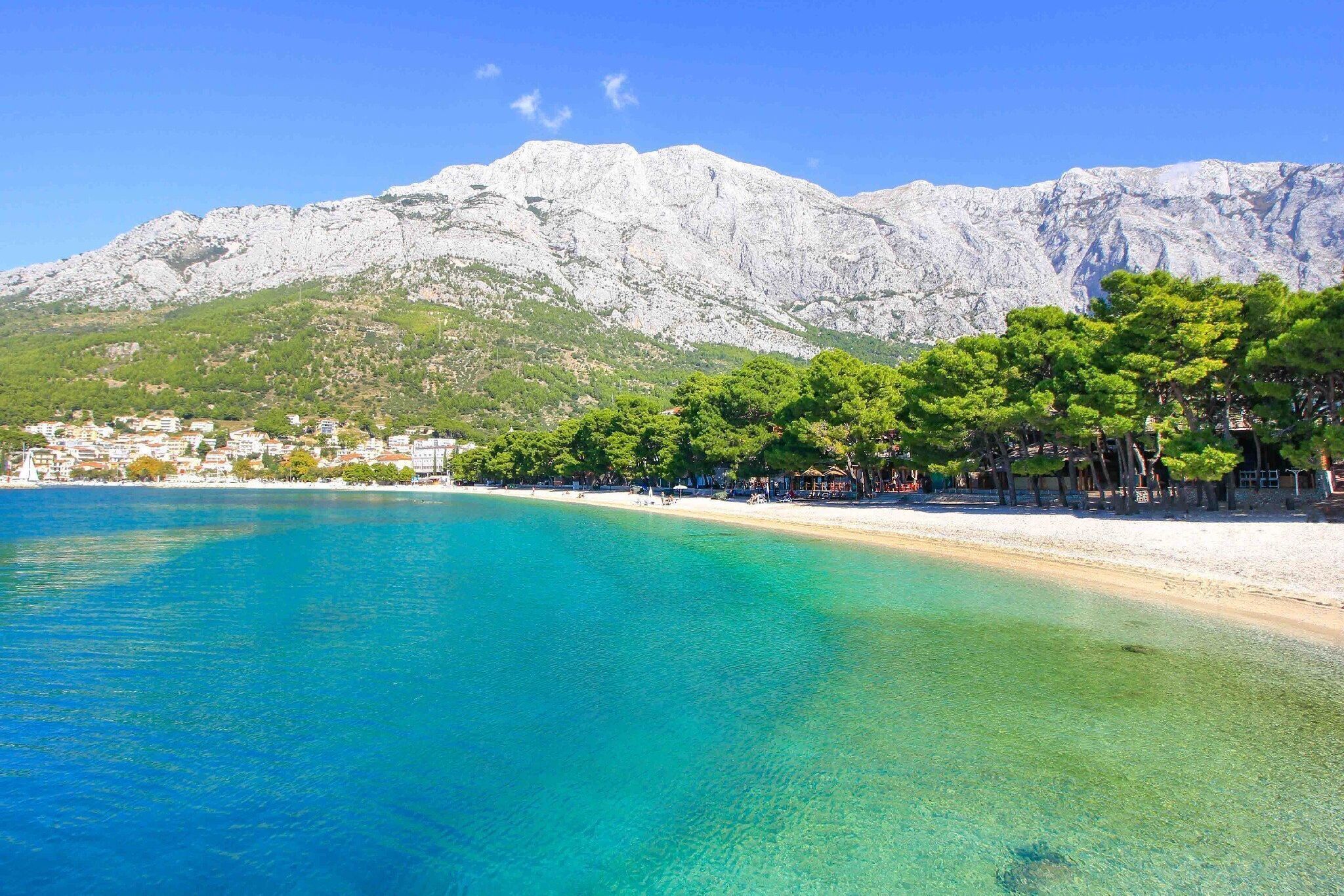 A coastal beach with clear turquoise waters, a sandy shore, and a backdrop of rocky mountains.