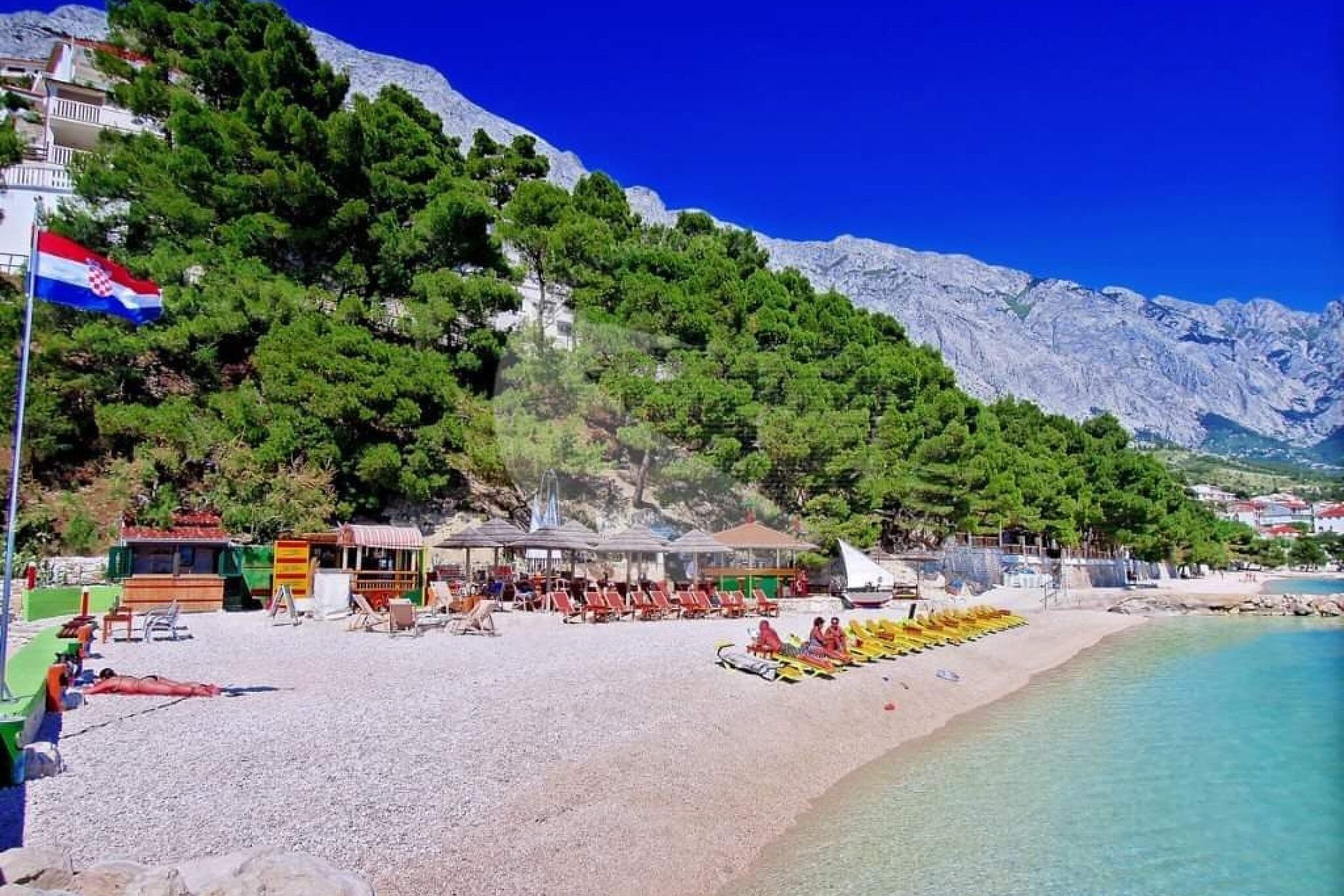 A beach with a flag, umbrellas, and beach chairs.
