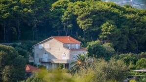 Une maison à deux étages, avec un toit recouvert de tuiles rouges, entourée d’arbres verdoyants et d’un paysage vallonné.