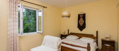A bedroom with a wooden bed, a small bedside table, a nightstand, a window with curtains, and a hanging decoration.