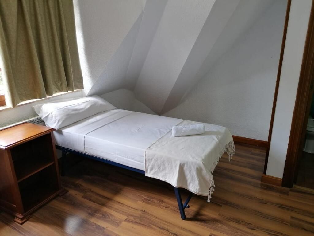 A small, single-bed room with a sloped ceiling, wooden flooring, a bedside table, and a window with curtains.