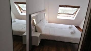 A small, single-bed room with a skylight and a window, wooden flooring, and a white bed.