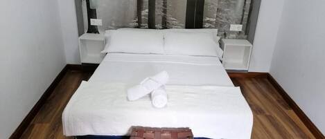 Un letto rifatto con una coperta bianca, una panca di legno ai piedi del letto e una testiera con motivo naturalistico.