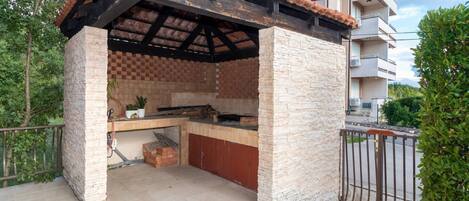 Un área de cocina al aire libre con pared de piedra y techo de baldosas, rodeada por una cerca y vegetación.