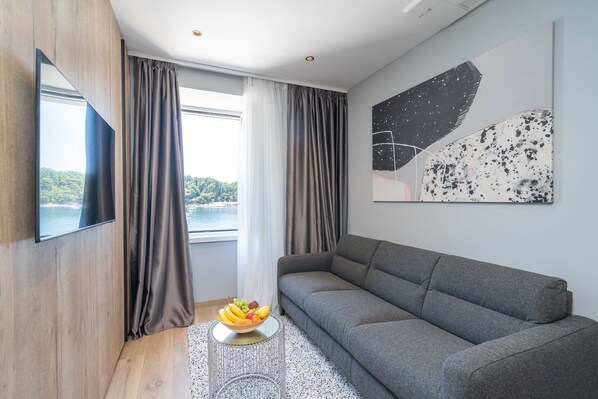 A modern living room with a grey sofa, a flat-screen TV mounted on a wooden wall, a window with curtains, and a fruit bowl on a small table.