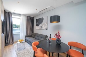 A modern living room with a grey sofa, orange chairs, a round black table with wine glasses and a bottle, a TV on the wall, and a window with curtains.