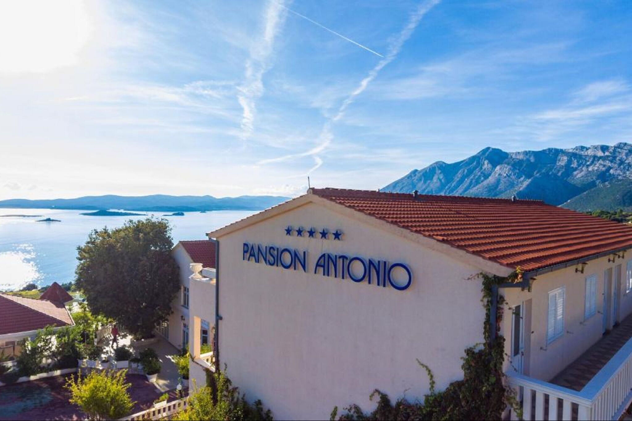 En byggnad med en skylt som visar texten ”Pensionat Antonio”, med utsikt över berg och en sjö eller ett annat vattendrag i bakgrunden.