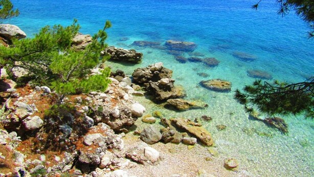 A coastal landscape with clear turquoise waters, rocky shore, and sparse vegetation.