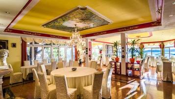 Un'ampia sala da pranzo con tavoli e sedie rotonde, un lampadario e grandi finestre che offrono vista sull'esterno.