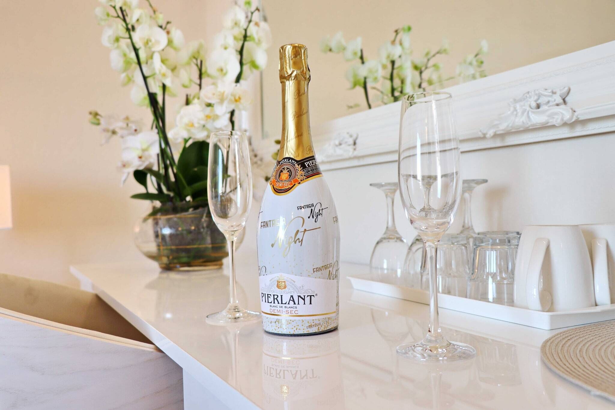Eine Flasche Pierlant Champagner, ein Glas und eine Vase mit weißen Blumen auf einem weißen Tisch.