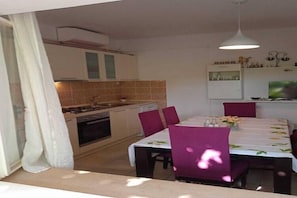 Un comedor con una mesa preparada para cuatro, una cocina al fondo y un televisor en la pared.