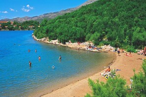 A beach with clear blue water, people swimming and sunbathing, and a forested hillside in the background.