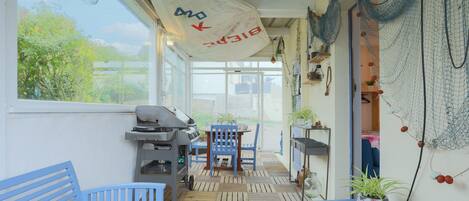 Ein überdachter Patio-Bereich mit Grill, blauen Stühlen und einem Tisch mit Topfpflanzen.