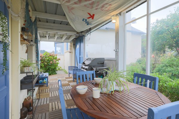 Une terrasse couverte avec une table en bois, des chaises bleues, un barbecue et des plantes en pot.