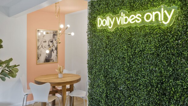 A modern dining area with a round wooden table, white chairs, a neon sign reading "Dolly vibes only", and a green leafy wall.