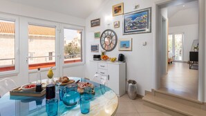 A modern dining area with a glass table, blue glassware, and a variety of food items. There are multiple windows providing natural light and views of the outside.