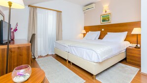 Habitación de hotel con cama doble, lámparas de noche, escritorio con lámpara, televisor, un jarrón pequeño con flores y un equipo de aire acondicionado.