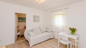 A minimalist room with a bed, a small table with a vase of flowers, and a window with sheer curtains. - Color Apartments - One Bedroom Apartment with Terrace and Partial Sea View (Blue) (Mlini)