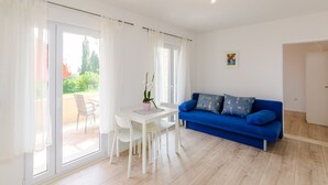 A bright living room with a white table, blue sofa, and a sliding glass door leading to an outdoor area. - Color Apartments - Standard One-Bedroom Apartment with Terrace and Partial Sea View (Red) (Mlini)