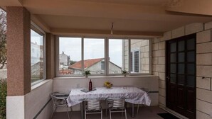 Un área de comedor al aire libre techada con mesa y sillas, ventanas amplias y vistas a edificios en el fondo.