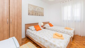 Una habitación con dos camas, un armario de madera, un cuadro en la pared y una ventana con cortinas traslúcidas.