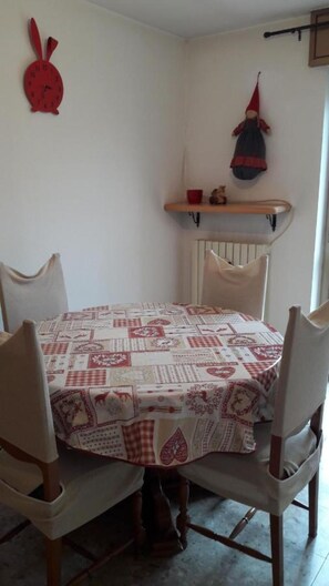 A dining table with a patterned tablecloth, four chairs, a red wall clock, and a shelf with a red hat and a small figurine.