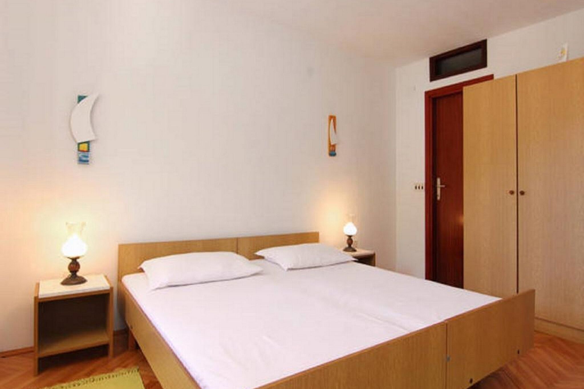 A single-bed room with a wooden headboard, two bedside lamps, a wooden wardrobe, and a small shelf.